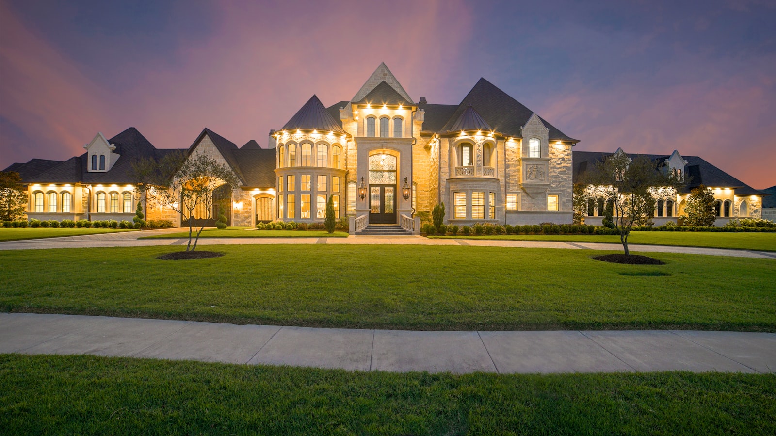 Grand estate home lit warmly at evening with manicured lawn and clean walkways